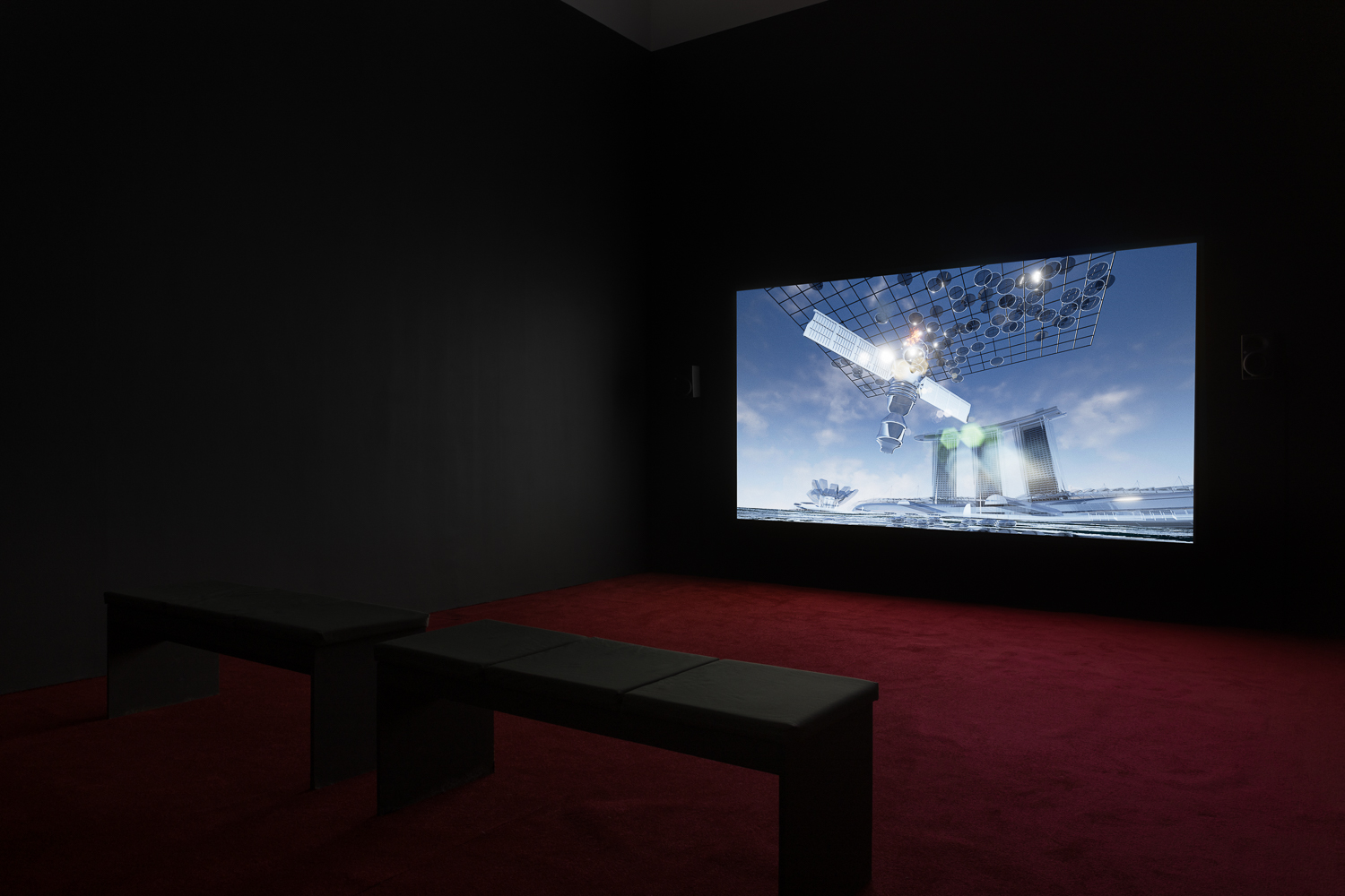 On the wall of a dark room is a projection of a CGI image of a satellite against a blue sky. The room has red carpet and a grey bench.