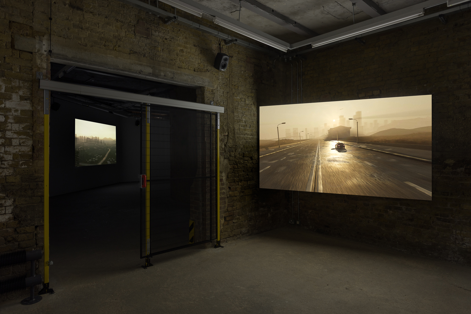 A projection of a CGI image of a car on a deserted road at sunrise or sunset in a brick walled gallery space. Over the gallery doorway is a yellow and black metal gate.