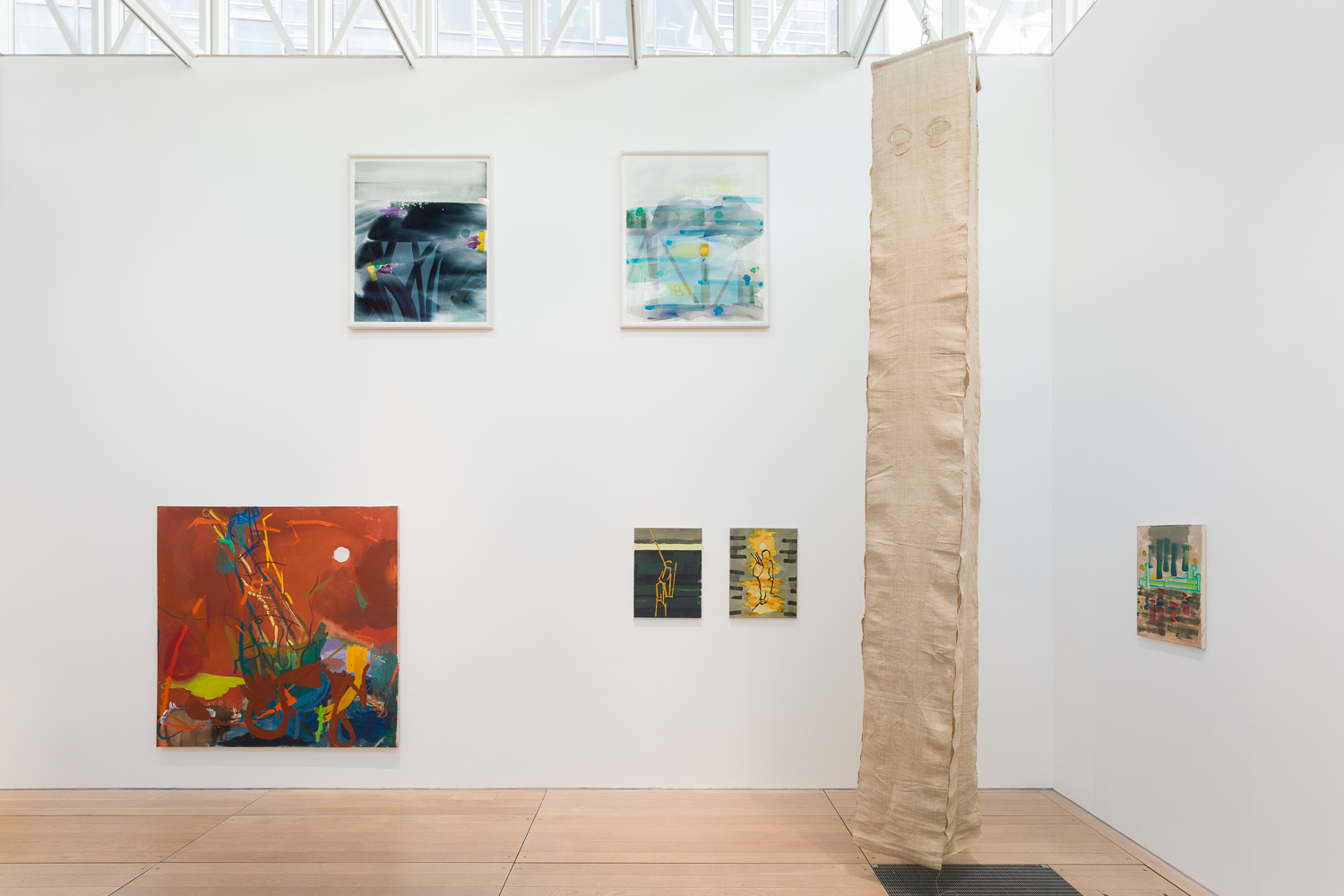 A white walled gallery with paintings hanging at varying heights on the walls. A long linen sculpture hangs from the ceiling in the corner the room.