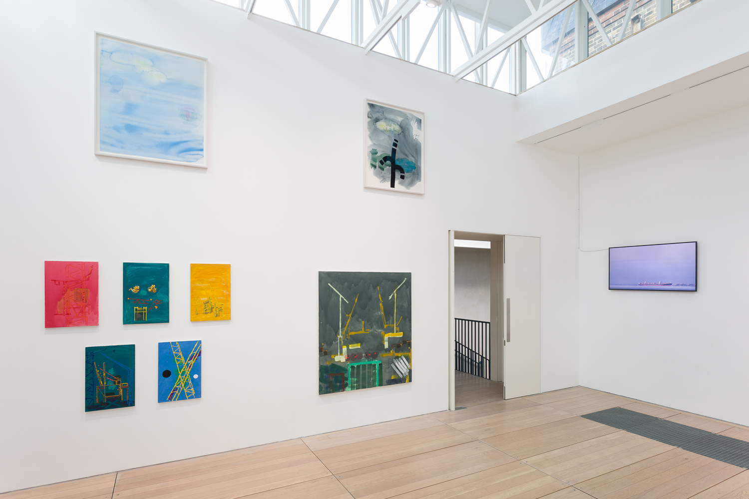 A white walled gallery with colourful abstract paintings hanging at varying heights on the wall. On the other wall hangs a flat screen monitor, showing the image of an industrial ship at sea.