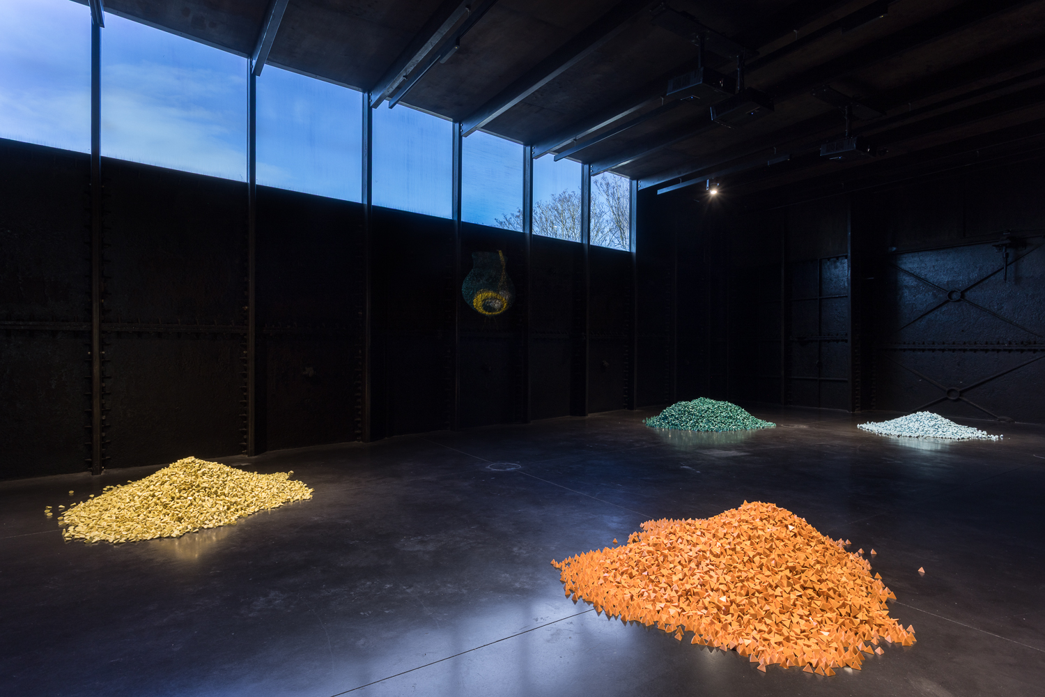 A black gallery space, previously an iron-lined Victorian water tank, with two piles of brightly coloured orange and yellow tumbling media on the floor.