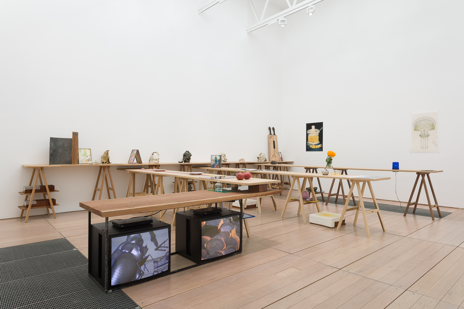 A white cube gallery space filled with trestle tables. Under one table is two monitors playing films. Others are topped with items such as pomegranates and a flower.