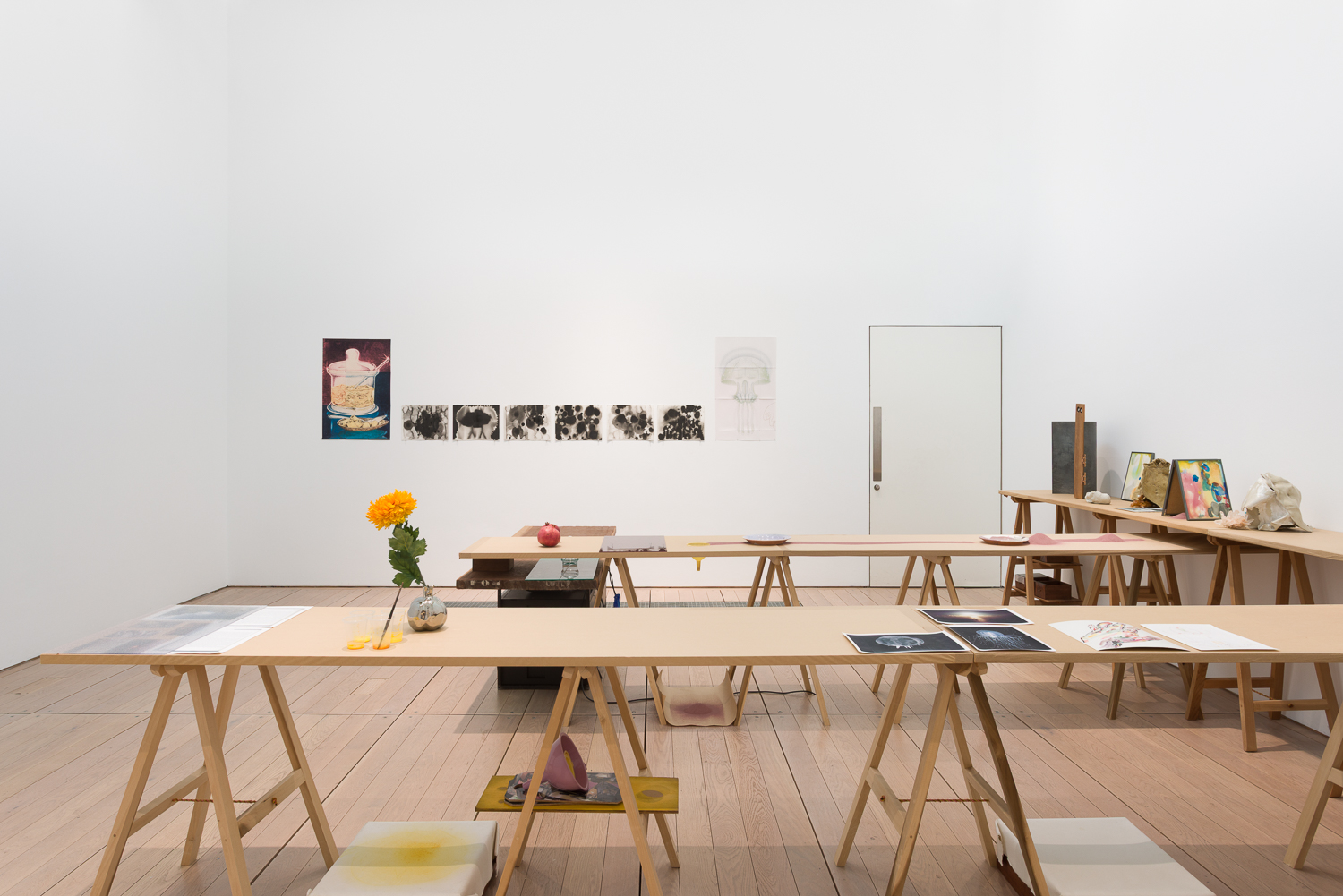 A white cube gallery space filled with trestle tables. The are topped with items such as paintings, printed matter, pomegranates and a flower. On the wall hang prints and watercolour paintings.
