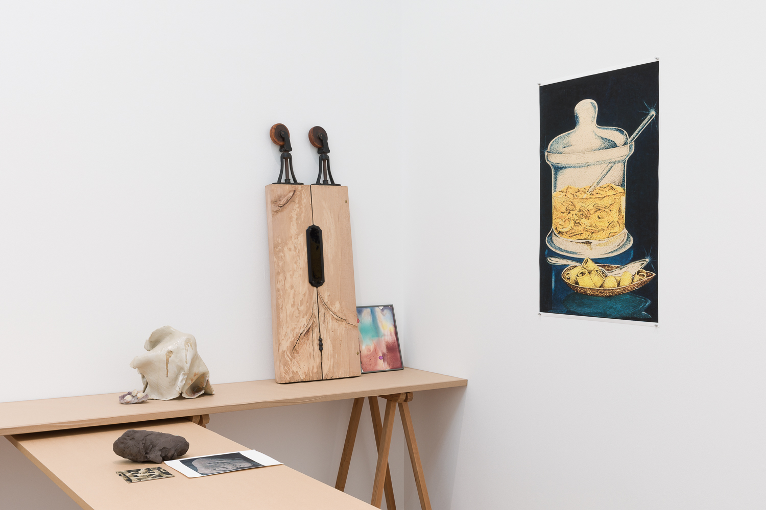 Trestle table with ceramic and wooden scultures, abstract watercolour paintings, and printed material on top. On the wall next to the table is an high contrast illustrated image of a glass jar.