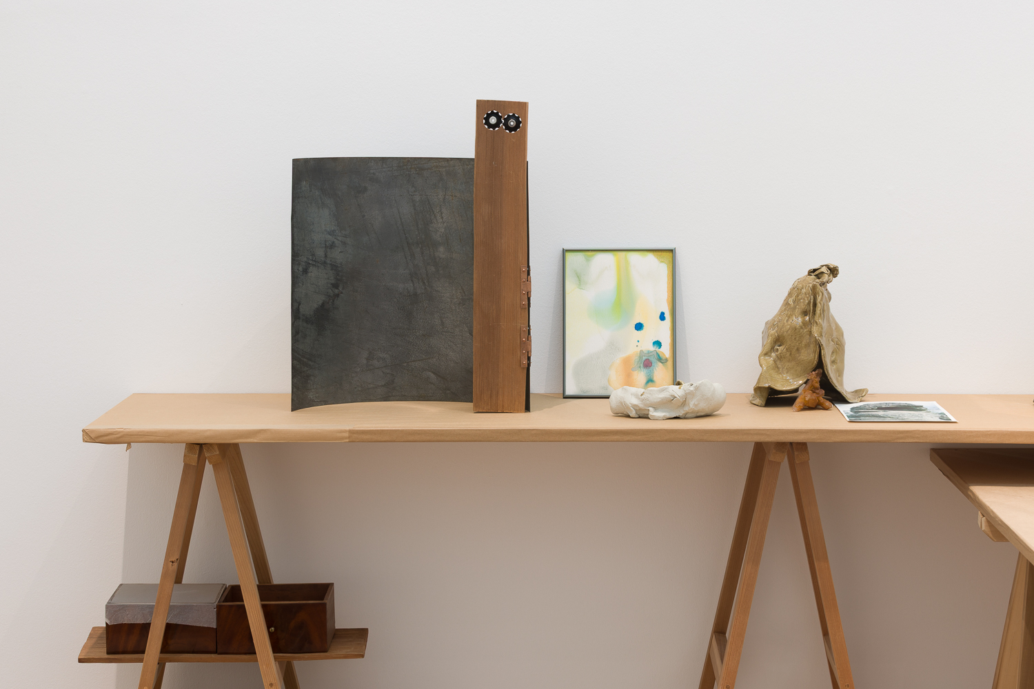 Trestle table with ceramic and wooden scultures, abstract watercolour paintings, and printed material on top.