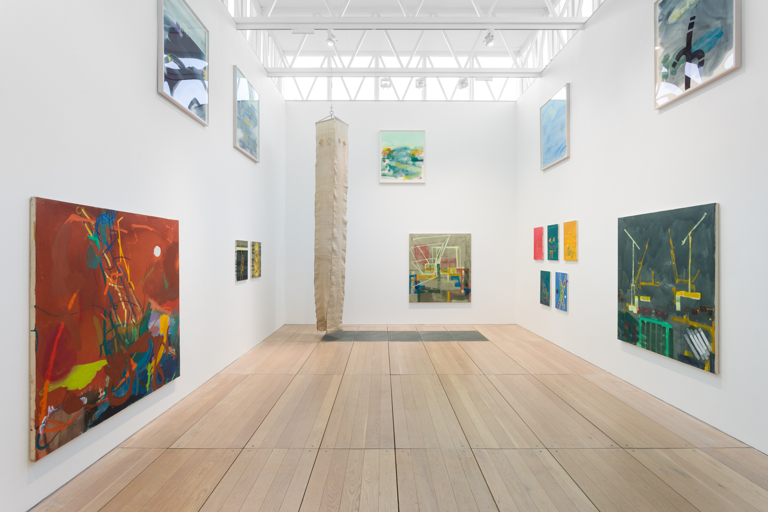 A white walled gallery with paintings hanging at varying heights on the walls. A long linen sculpture hangs from the ceiling at the rear of the room.
