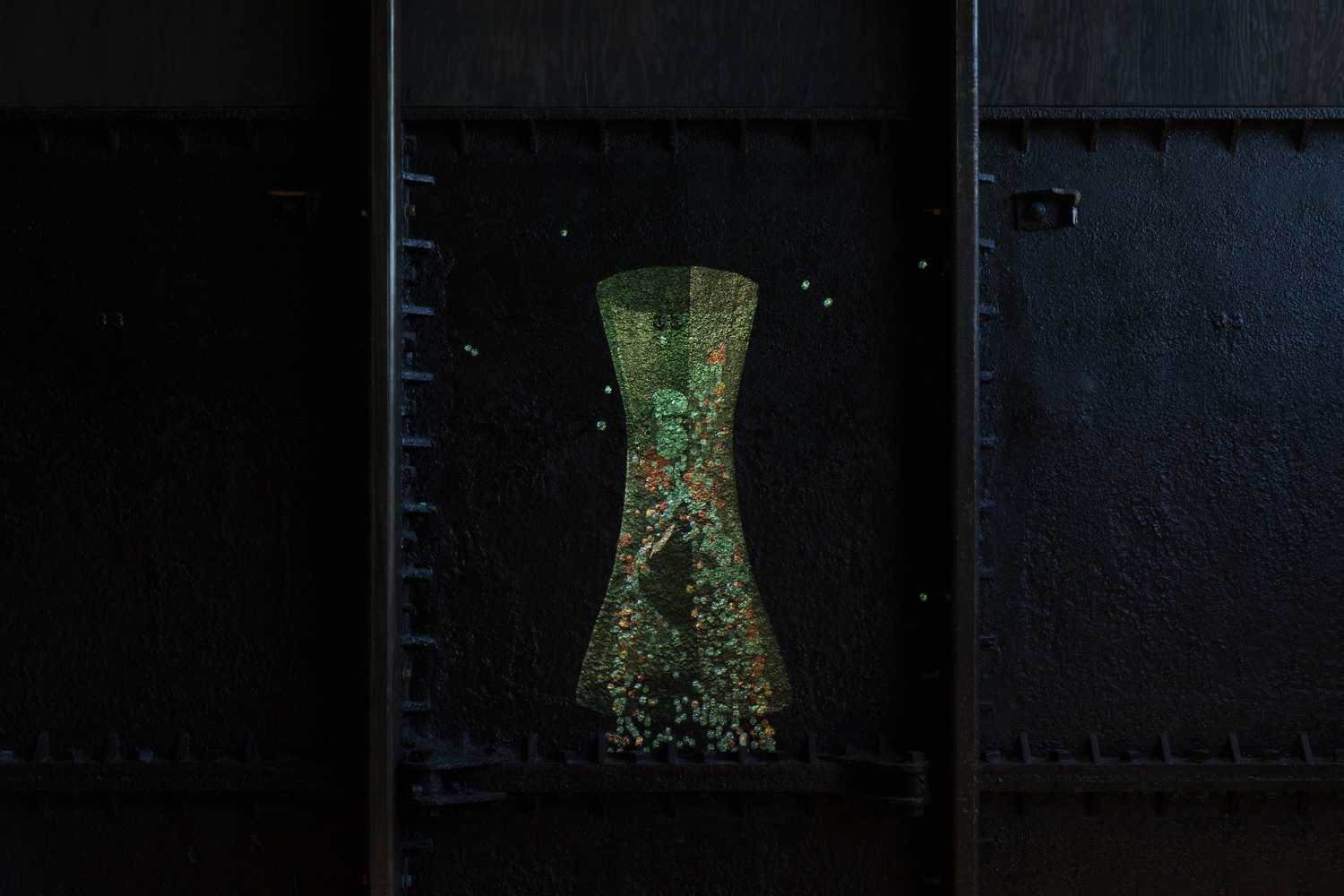 A textured black iron wall on which a projected animation shows balls falling through a green funnel, used to model the behaviour of people in crowds.