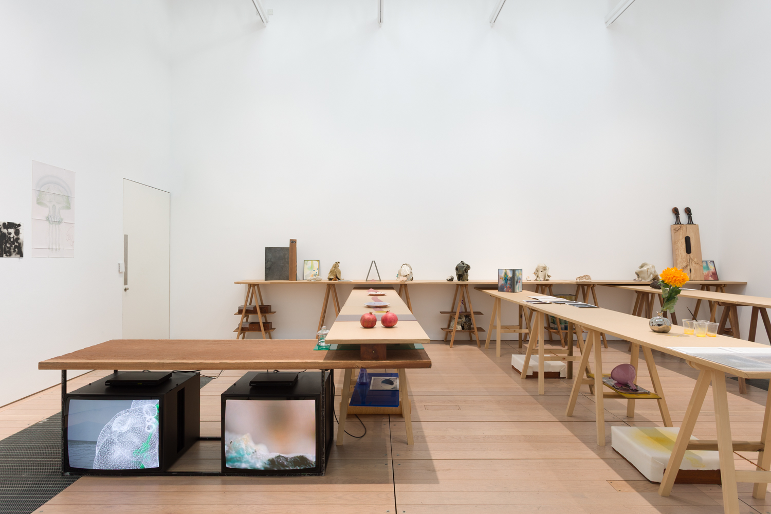 A white cube gallery space filled with trestle tables. Under one table is two monitors playing films. Others are topped with items such as pomegranates and a flower.