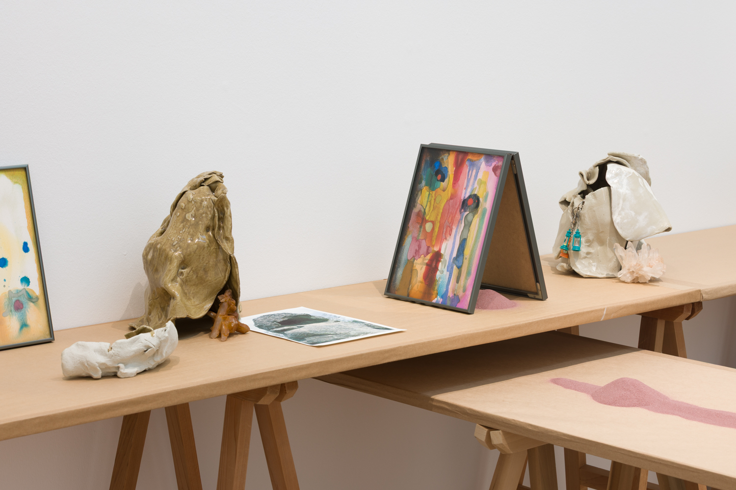 Trestle table with ceramic sculptures, abstract watercolour paintings, and a trail of lilac sand on top.