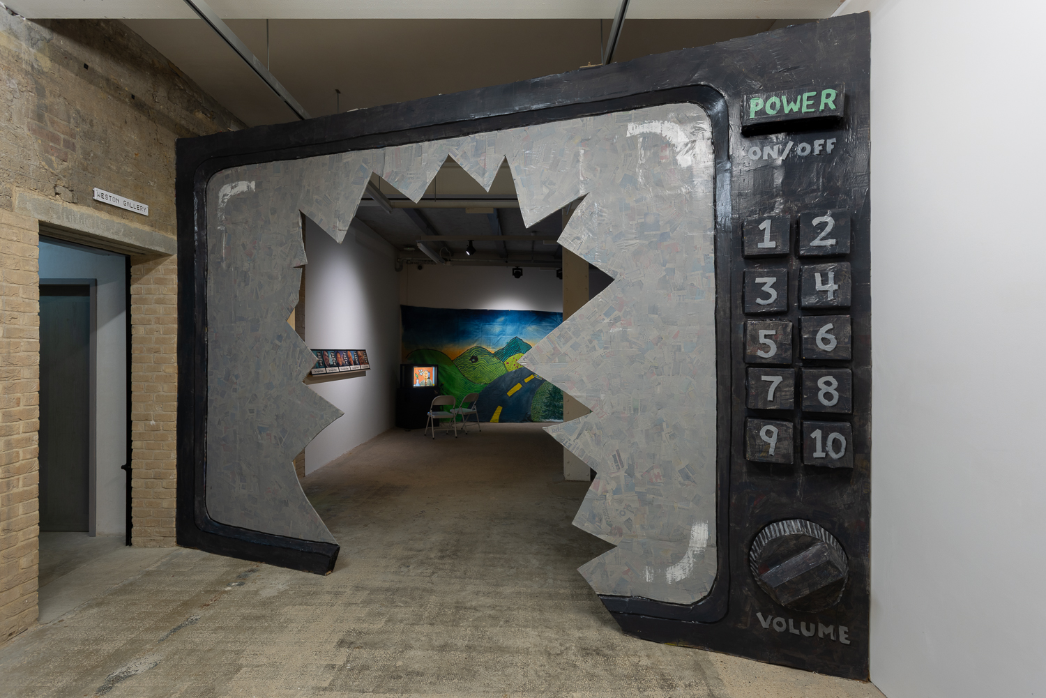 A large hand painted television, made of cardboard and papier-mâché, with a hole in the middle resembling a smashed screen. The hole forms a doorway, though which there is an art gallery exhibition.