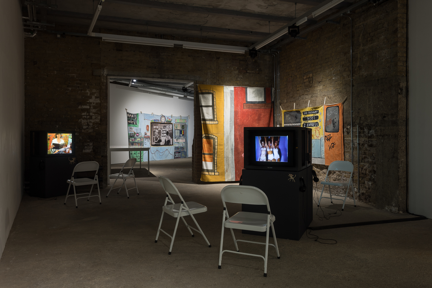 Brick walled gallery containing television monitors on dark grey plinths, with two white chairs in front of each. Colourful hand painted backdrops hang on the walls.