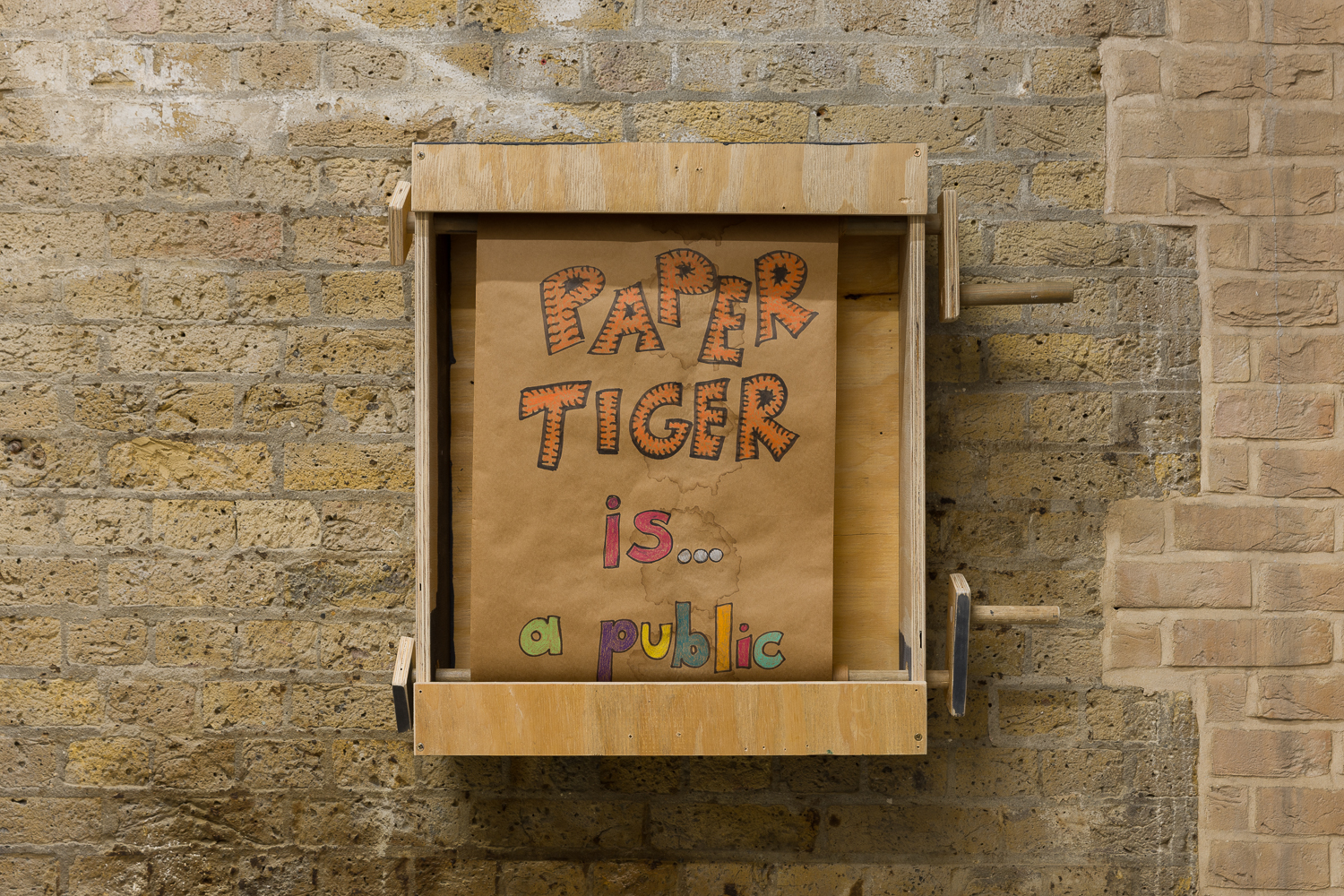 Wooden box with two crank handles hangs on an exposed brick wall. In the box is a roll of paper with colourful hand written text which reads 'PAPER TIGER is a public...'