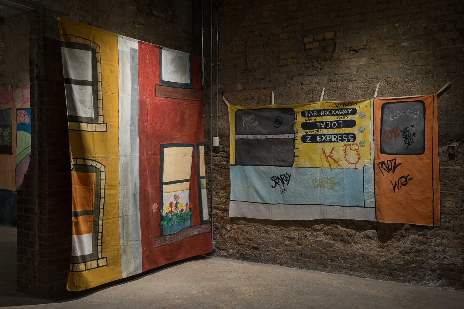 Exposed brick walls with two colourful hand painted backdrops hanging on them. One depicts a graffitied subway cart, and the other the facade of a red brick house.