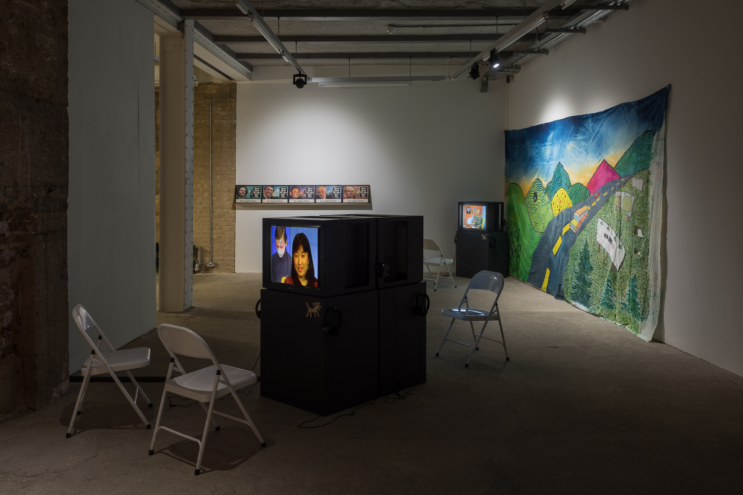 A white walled gallery containing television monitors on dark grey plinths, with two white chairs in front of each. A colourful hand painted backdrop hangs on the wall.