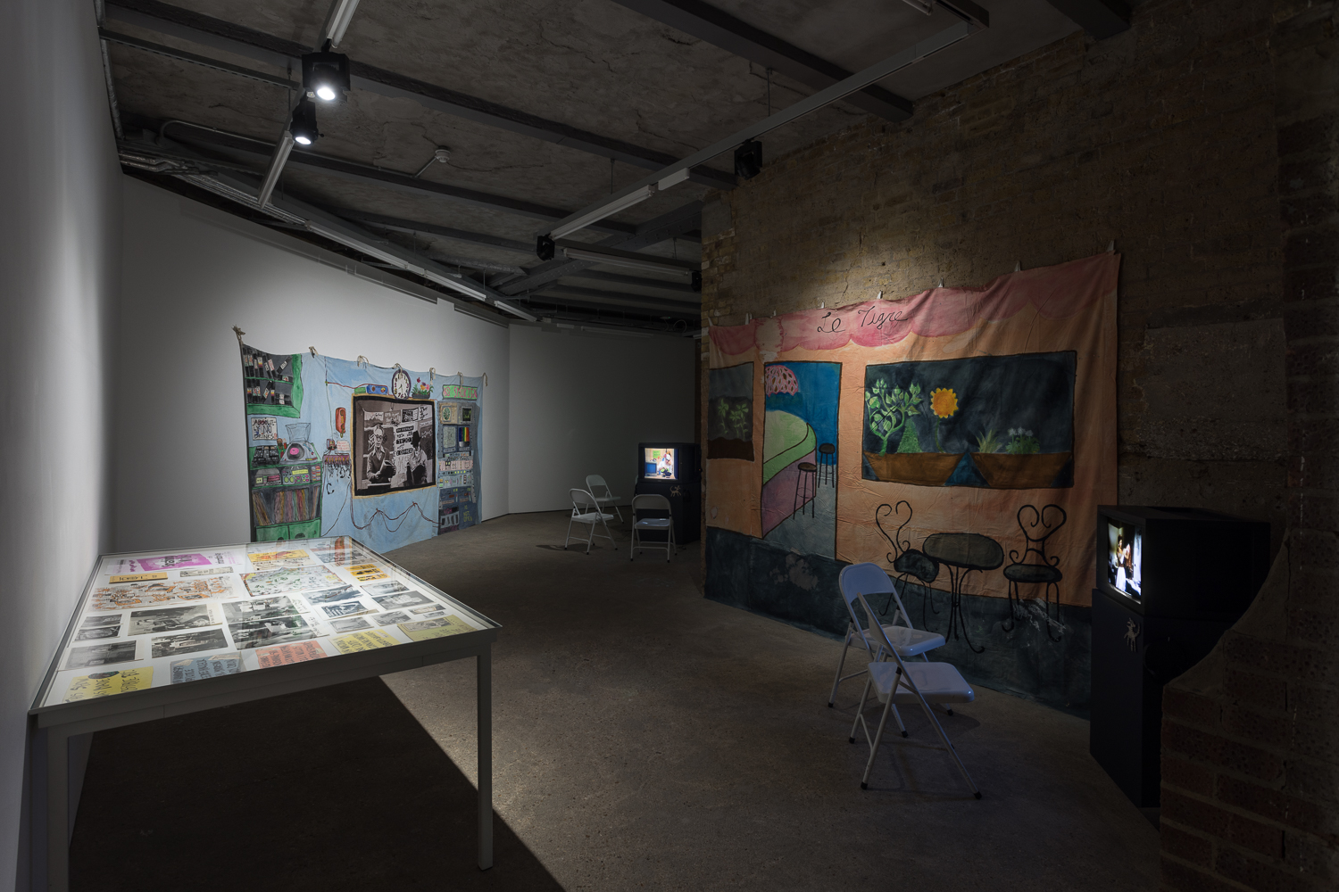 A gallery containing television monitors on dark grey plinths, with two white chairs in front of each. Colourful hand painted backdrops hang on the walls. A vitrine containing archival materials is in the foreground.