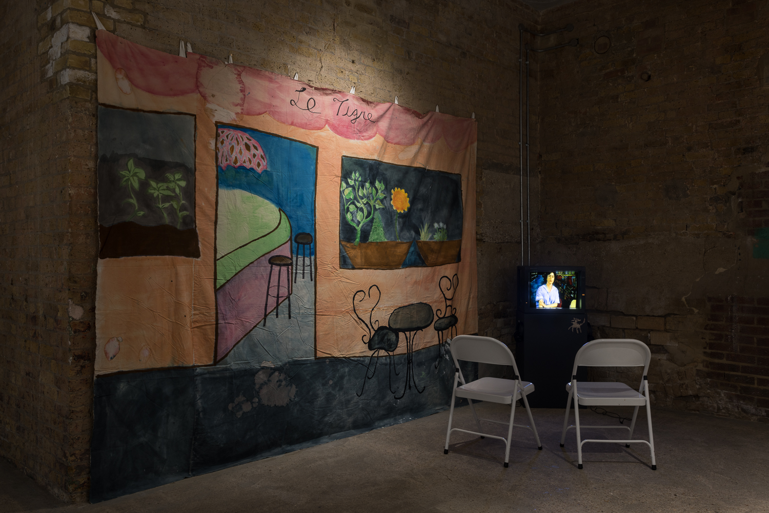 Brick walled gallery containing a television monitor on a dark grey plinth, with two white chairs in front. On the wall hangs a hand painted backdrops depicting a cafe front.