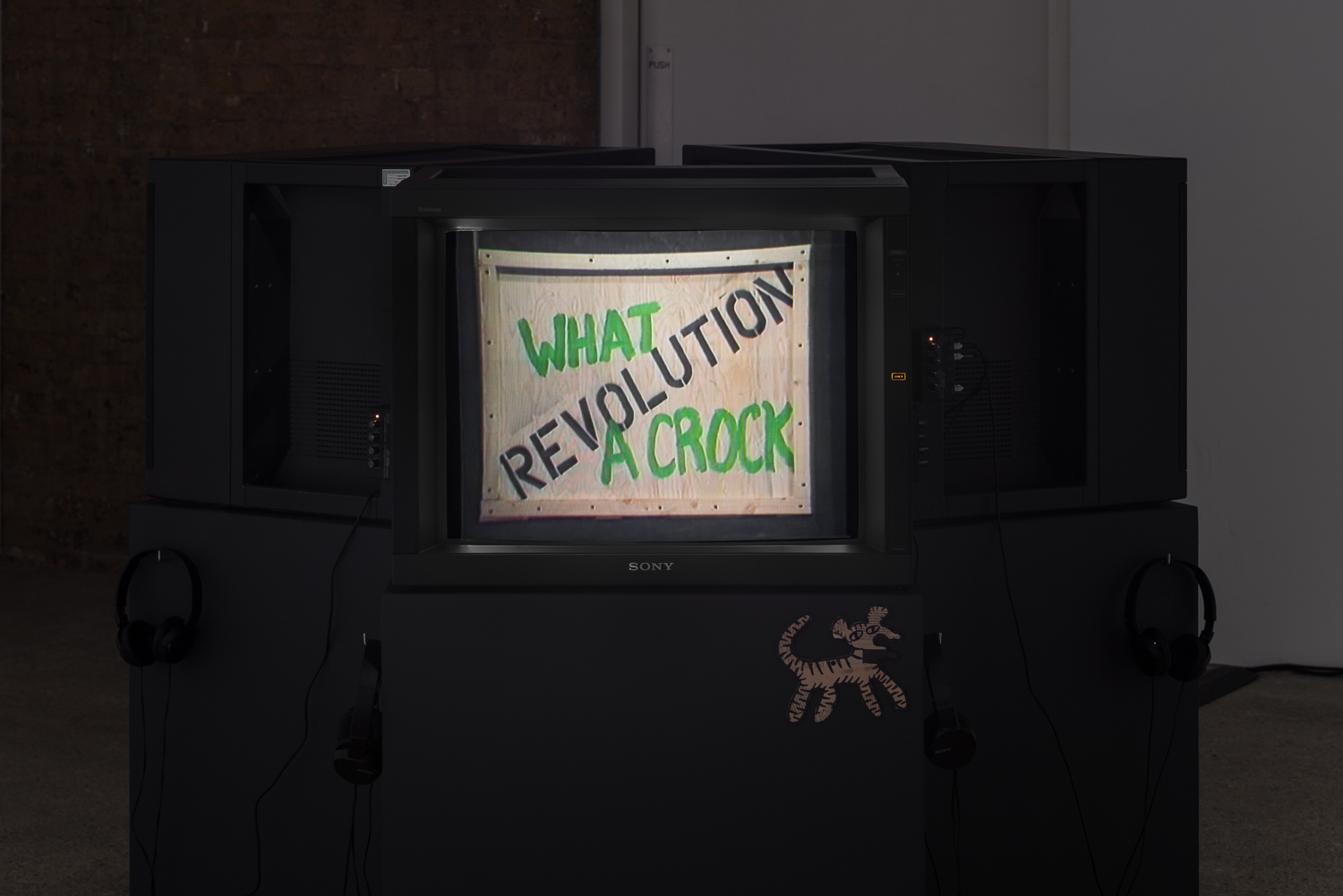 A television monitor on a dark grey plinth displays the image of a box. On the box is spray painted 'REVOLUTION WHAT A CROCK'.