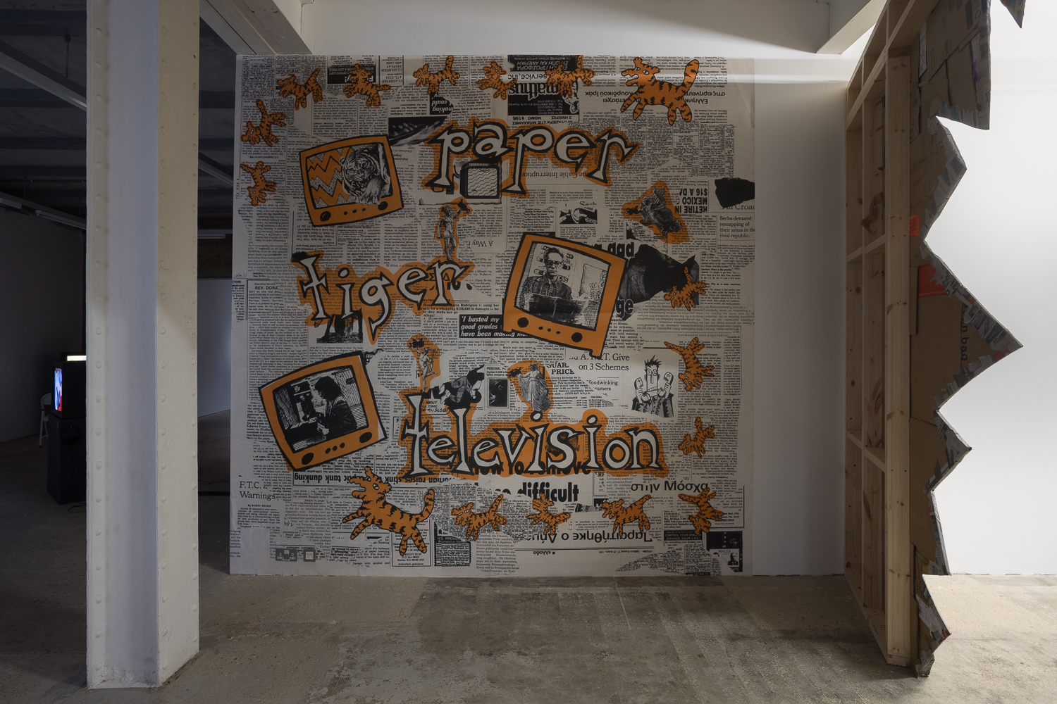 A gallery wall is wallpapered with a collaged image of newspaper cuttings. On top in large orange letters reads 'Paper Tiger Television'.