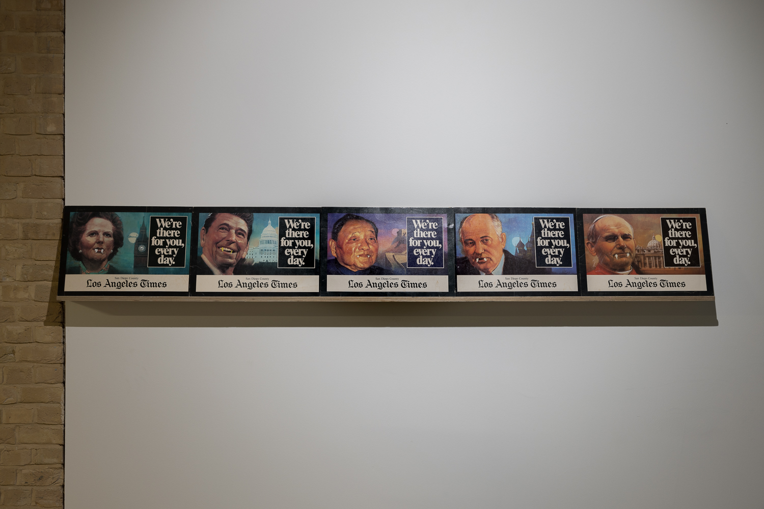 On a shelf on a gallery wall sit 6 poster images advertising the Los Angeles Times, displaying portraits of famous politicians. The images have been altered with white correction fluid to give the faces vampire fangs.