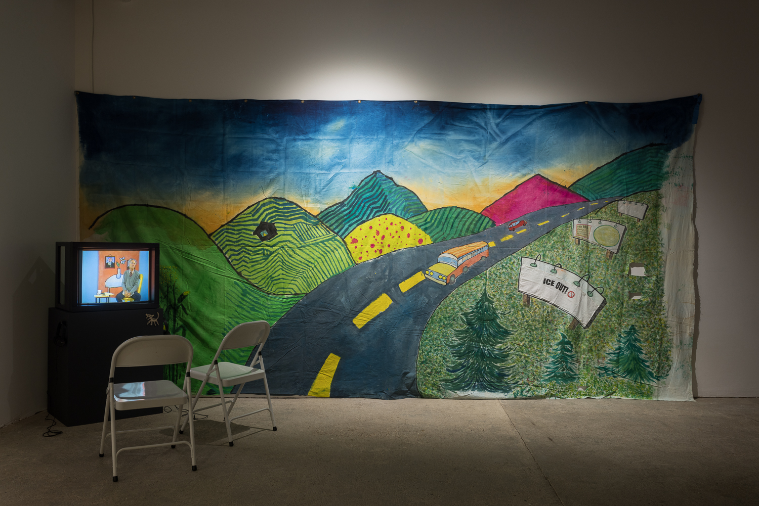 A large colourful hand painted backdrop depicting a road through a hilly landscape hangs on the wall. In front of it is a television monitor on a dark grey plinth, with two white chairs in front.