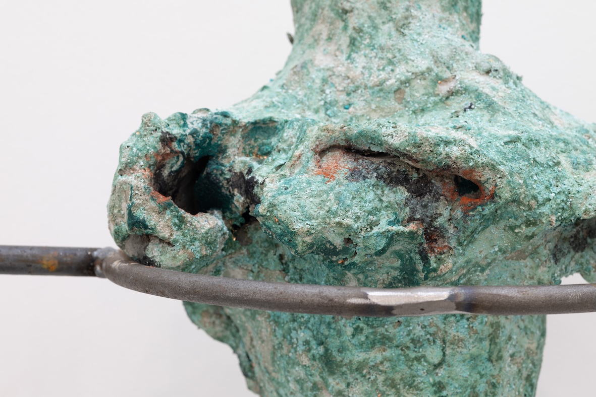 A metal structure supports a verdigris colored organic shaped sculpture. There are hollows in the form, it tapers at the top and bottom.