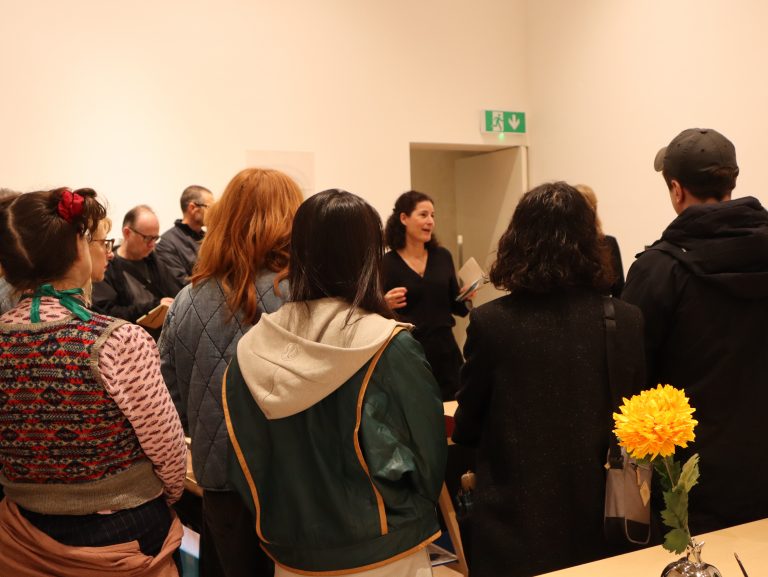 A group of people gathers in a room, listening to a woman speaking. They wear casual clothing, and the mood is attentive. A vase with a yellow flower is in the foreground.