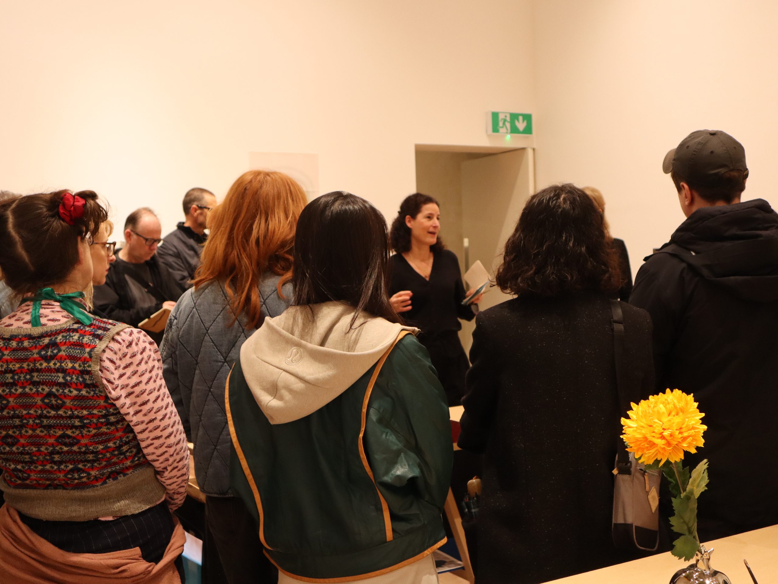 A group of people gathers in a room, listening to a woman speaking. They wear casual clothing, and the mood is attentive. A vase with a yellow flower is in the foreground.