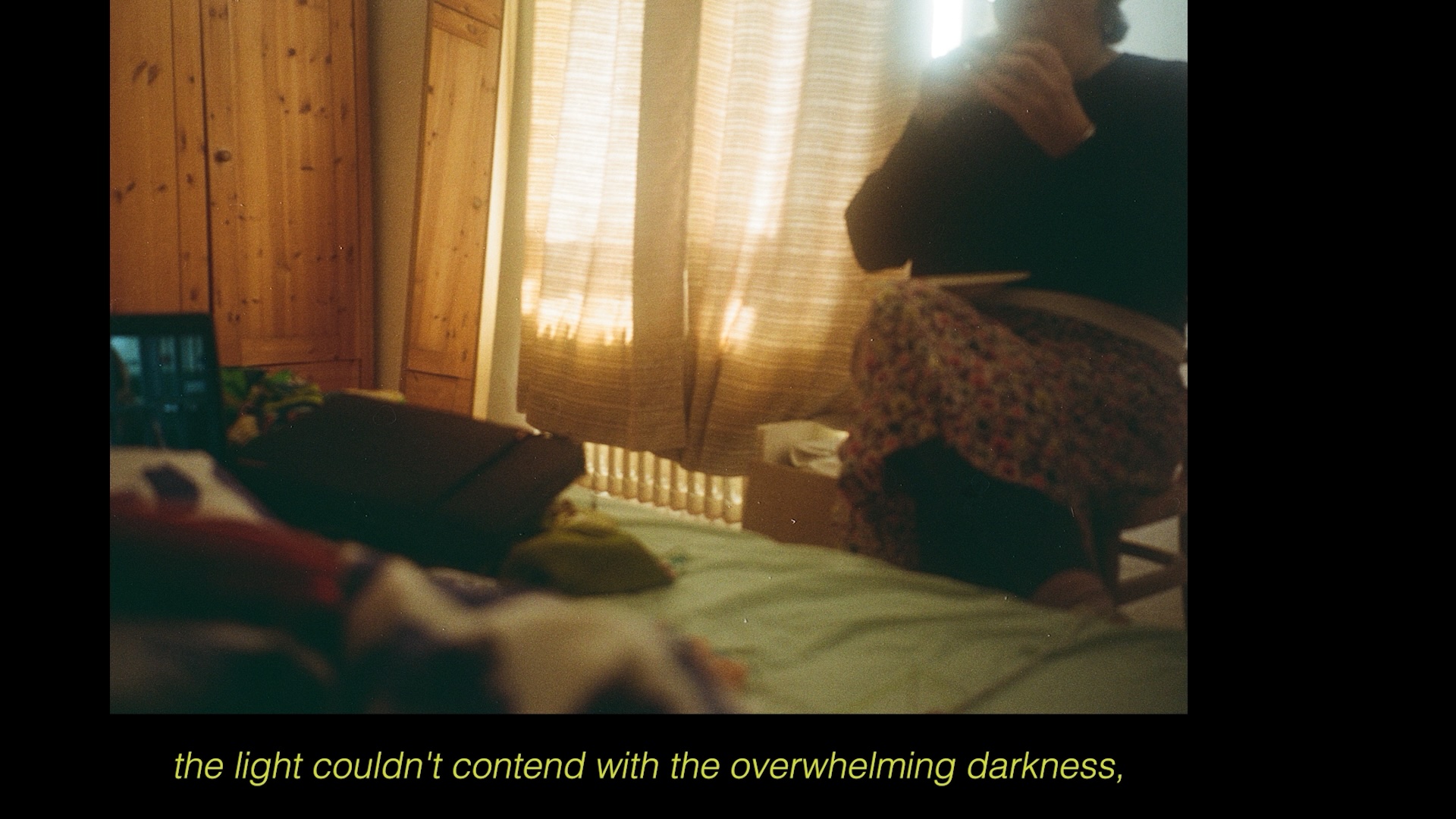 A photograph showing a dimly lit room shows a woman sat beside a bed holding up a phone camera. The figures face is obscured. They sit in front of drawn curtains, which light glows through. A cupboard is in the corner. Text below on a black backdrop reads "the light couldn't contend with the overwhelming darkness".