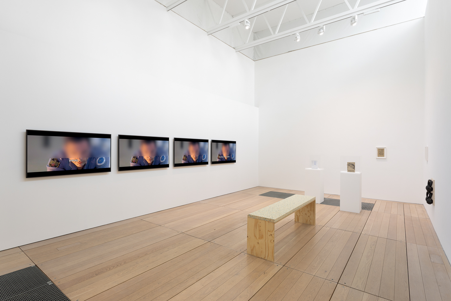 a white cube gallery with four identical screens on the left showing the same image. A wooden bench is placed in front and in the back of the image we see two plinths, each displaying a small rectangular artwork. a small rectangular artwork hangs on the wall behind.