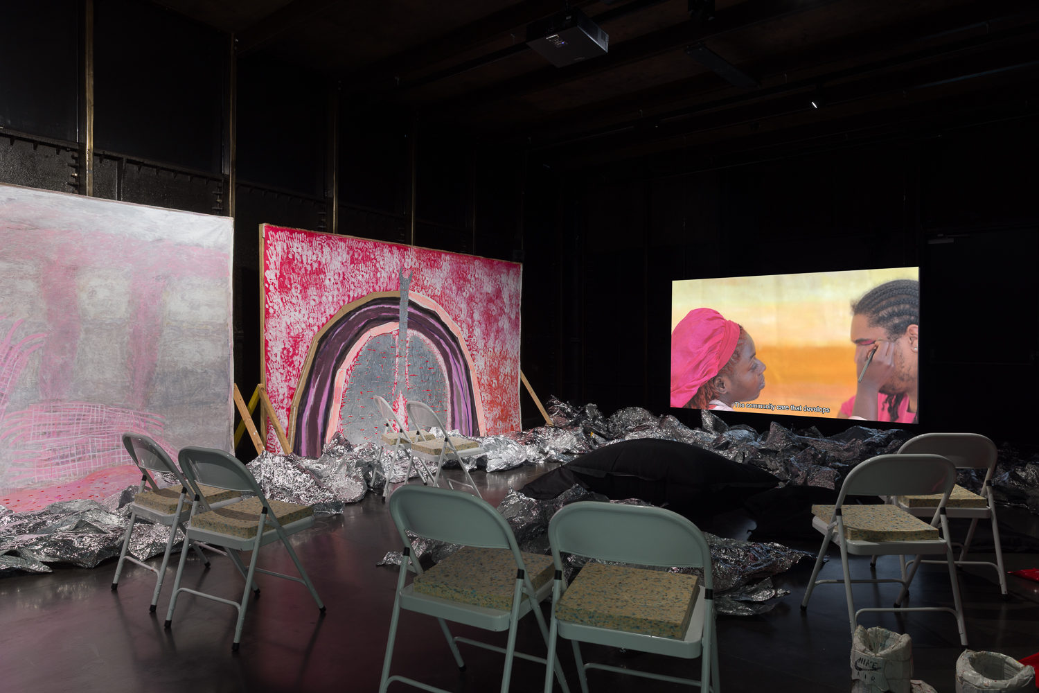 a dark gallery space, to the left are two large paintings stretched over wooden structures. Both are abstract and pink. In the background on the left is a large screen showing a film. In front of this is lots of silver foil and white chairs with square cushions on them.