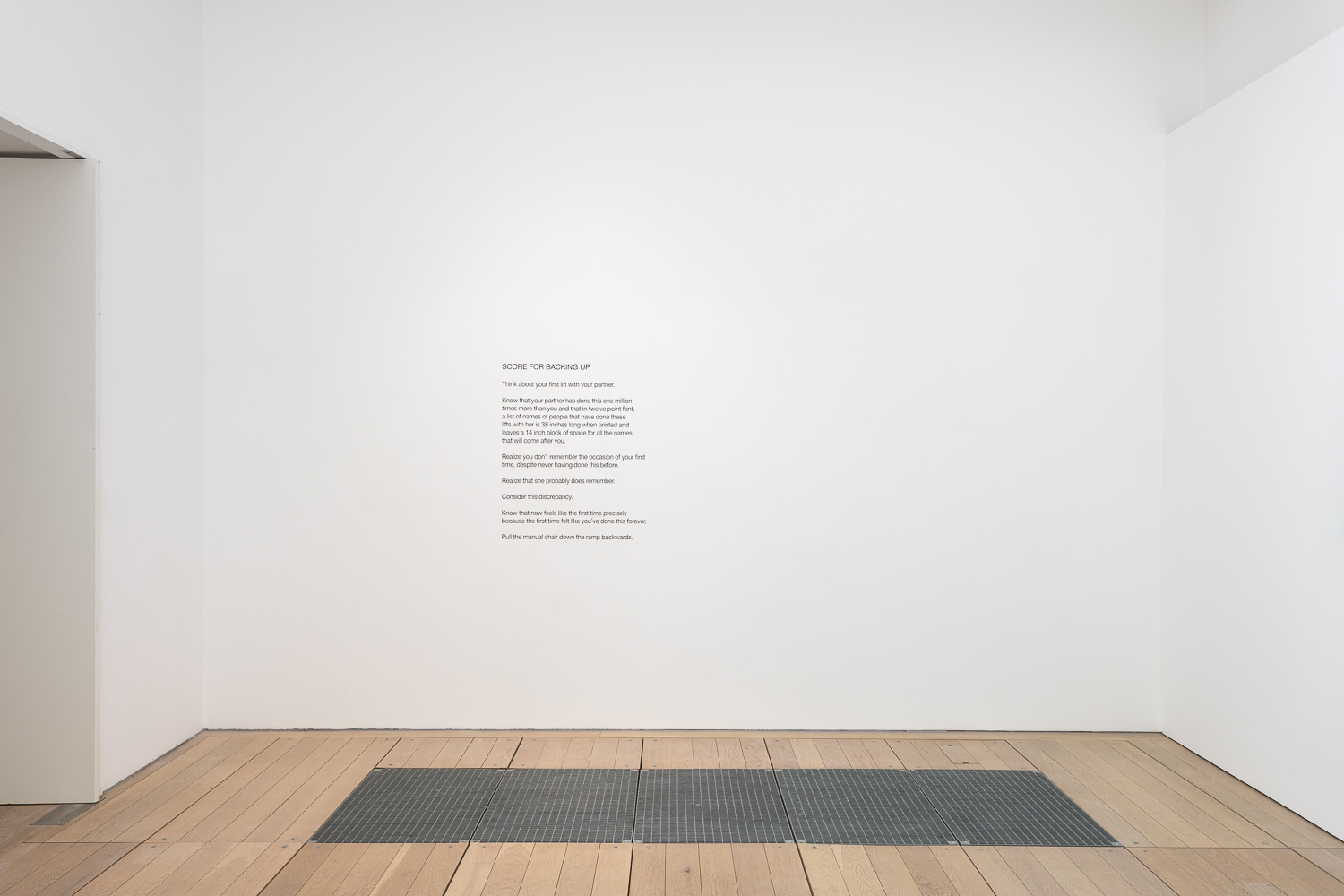 a white cube gallery with a poem in black font neatly on the wall slightly off centre to the left. a wooden floor has a metal grate in it.
