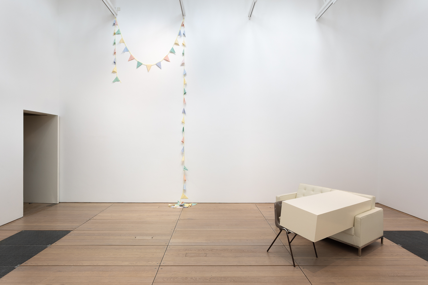 a white cube gallery. On the right is an art installation in which a white plinth is laid across a brown chair and a white sofa. In the background to the left a pastel coloured line of bunting is hung from the ceiling, draped against the back wall.