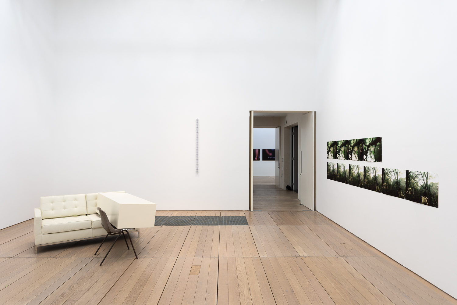 a white cube gallery with an art installation to the left encompassing a white sofa and brown four legged chair that a white plinth is placed across. on the right wall are a series of green-coloured photographs lined up one next to the other in two rows.