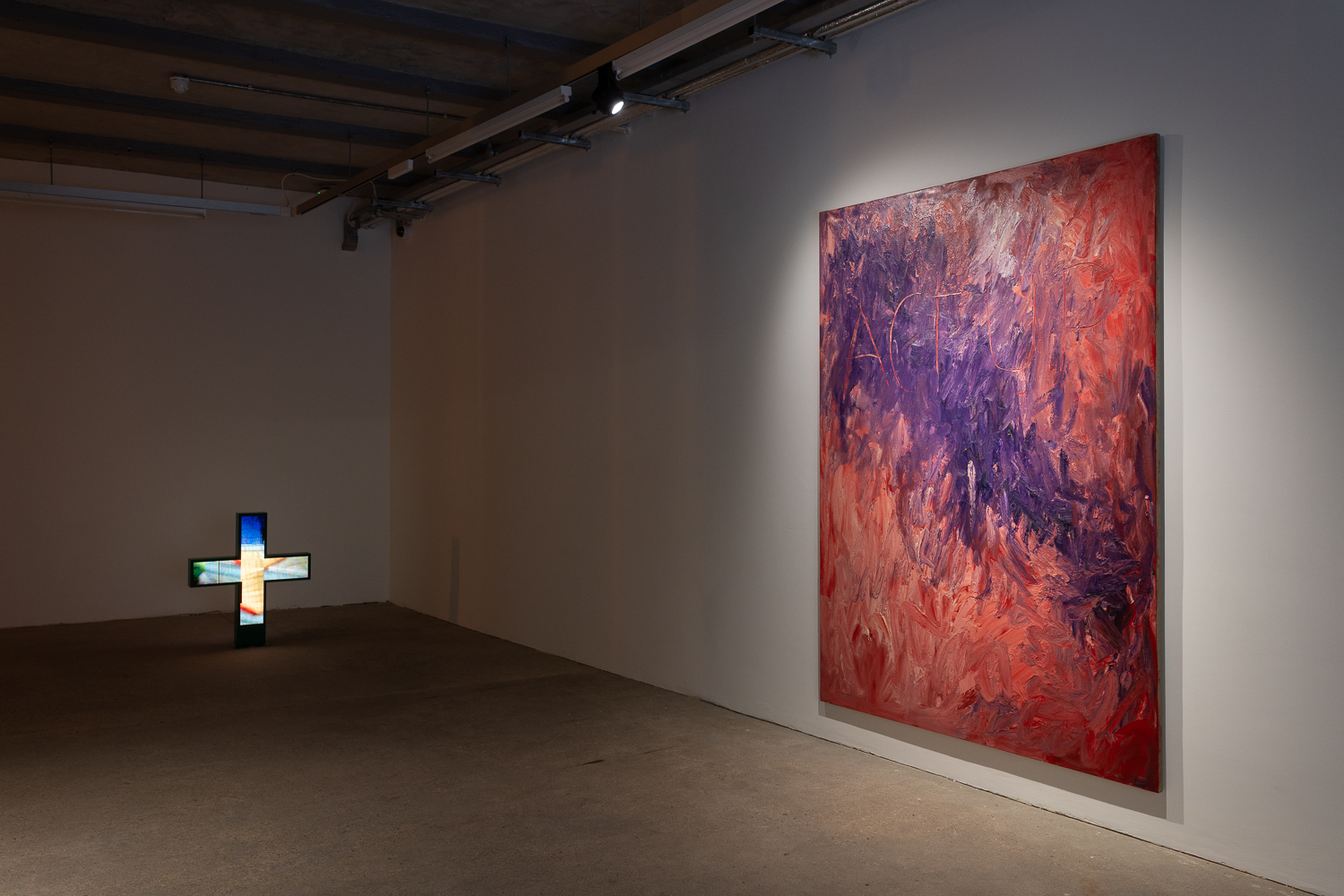 An installation in a gallery with a large purple and pink abstract painting hanging on a white wall to the right. In the background a pharmacy cross glows different colours.