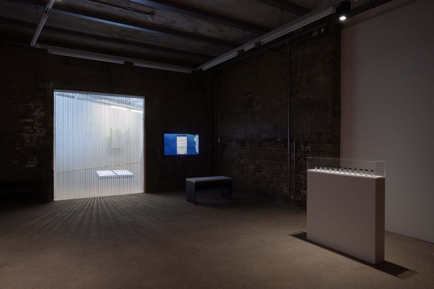 an installation in an art gallery. On the left a beaded curtain hangs in a doorway. To its right is a television screen showing a film with a bench in front of it. To the right in the foreground is a wide, but narrow plinth holding a display of tiny jars.