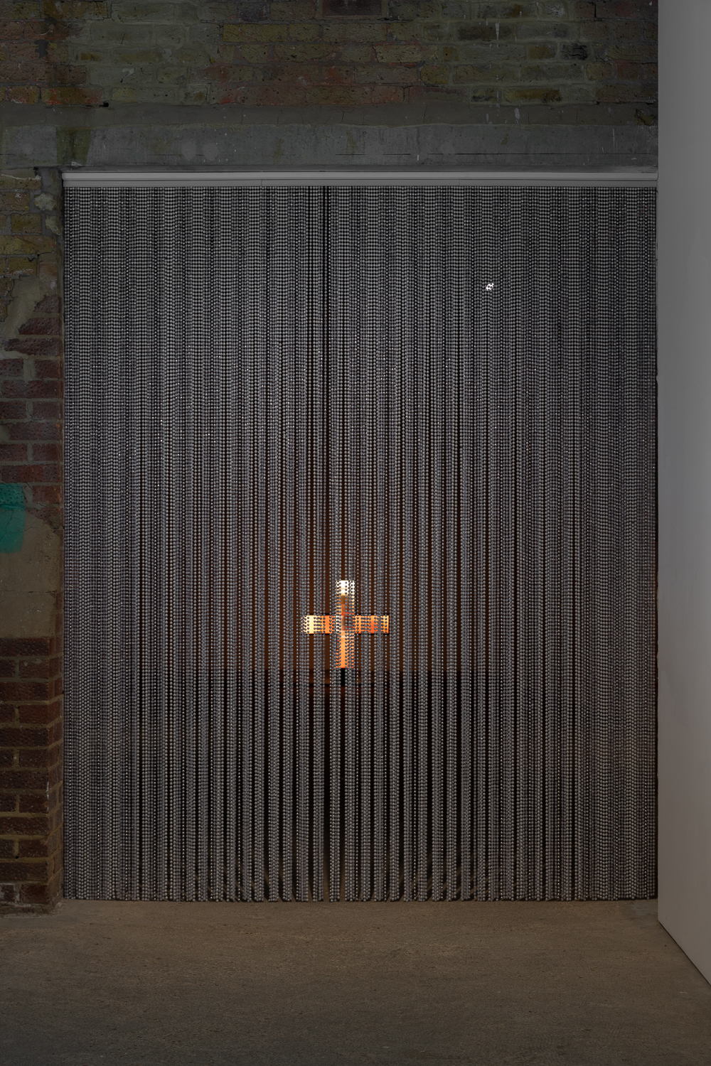 a beaded curtain hanging in a wide doorway. a brick wall surrounds it and through the curtain we see an illuminated pharmacy cross, glowing orange.