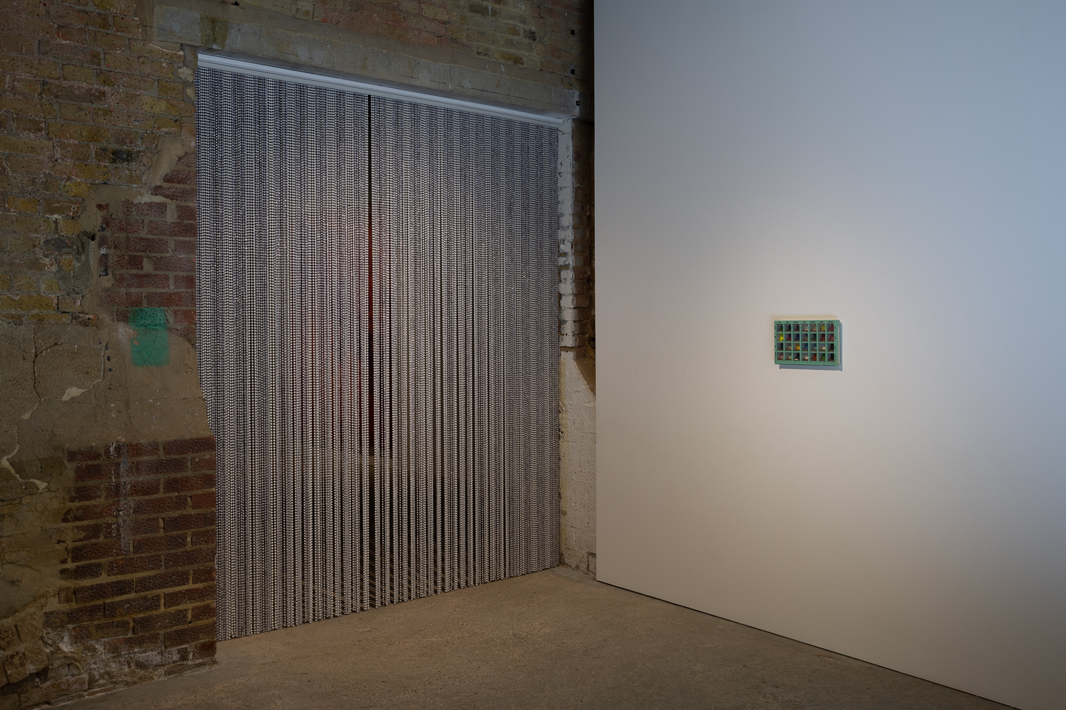 an installation in an art gallery, consisting of a beaded curtain hanging in a wide doorway in a brick wall. To its right hanging on a white wall is a small pale blue box with gridded windows in it.