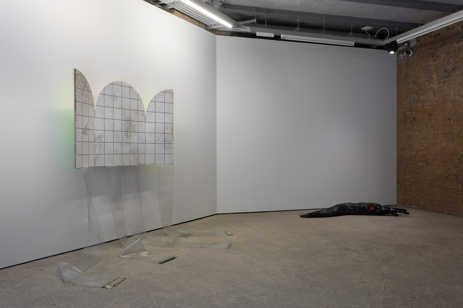 An installation inside of a white walled gallery, with a brick wall to the right side. A triptych painting showing a grid hangs on the left wall, with plastic strips trailing off it. A giant slug lies on the floor to the right.