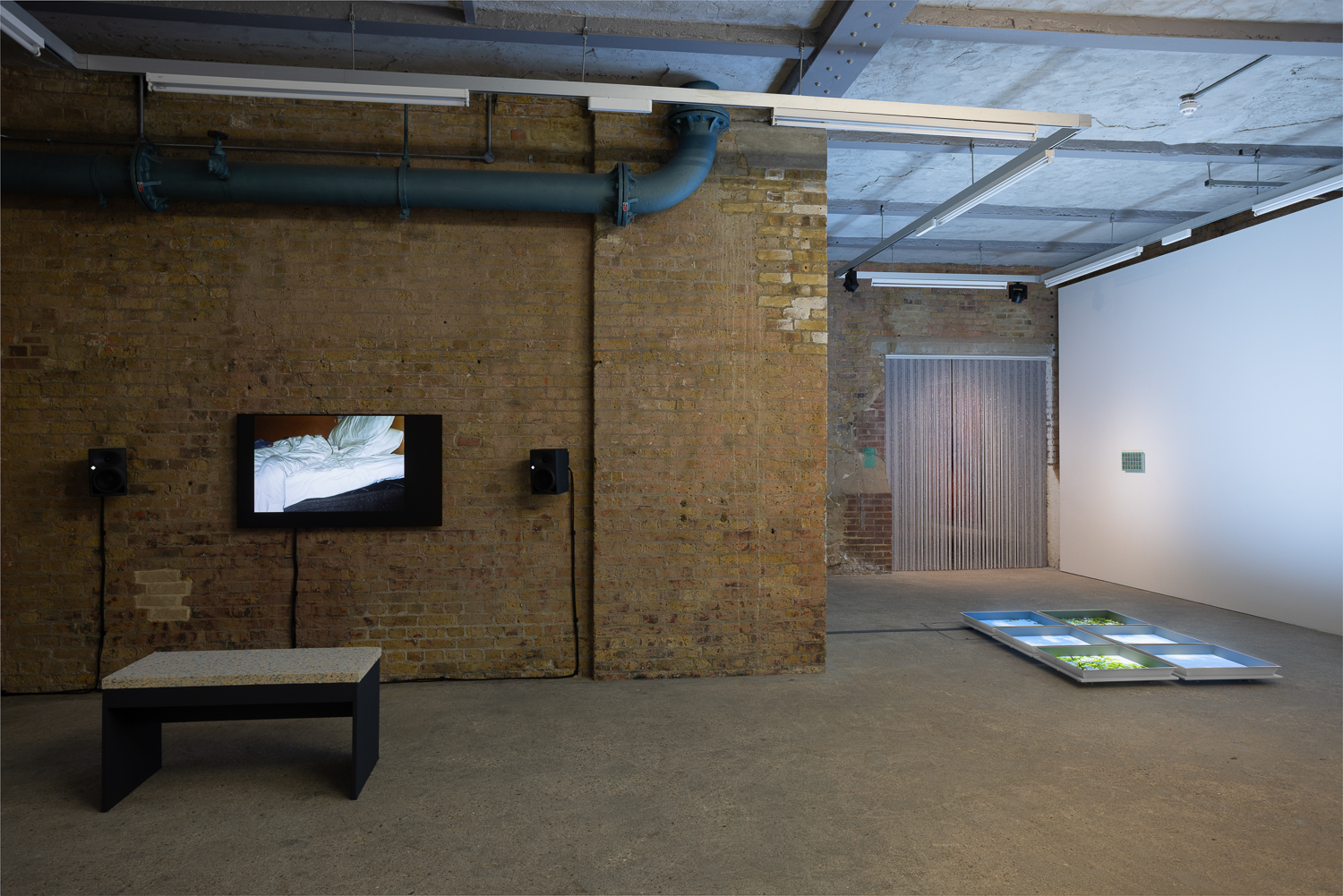 An installation shot showing a gallery with brick and white walls. On the left brick wall a tv screen is installed showing a film, with speakers either side and a bench in front. To the right a set of six square screens lie on the floor showing a green/blue image that isn't quite visible at this angle. A beaded curtain hangs in the doorway in the background.