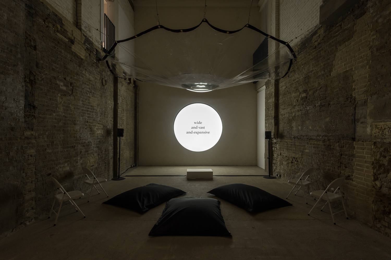 an art installation in a brick walled room with a high ceiling. at the back of the room is a circular screen glowing bright white with slightly imperceptible text written on it. hanging above the room is a circular, plastic tarpaulin with a little pool of water gathered in its middle. three large black beanbags are placed on the floor.