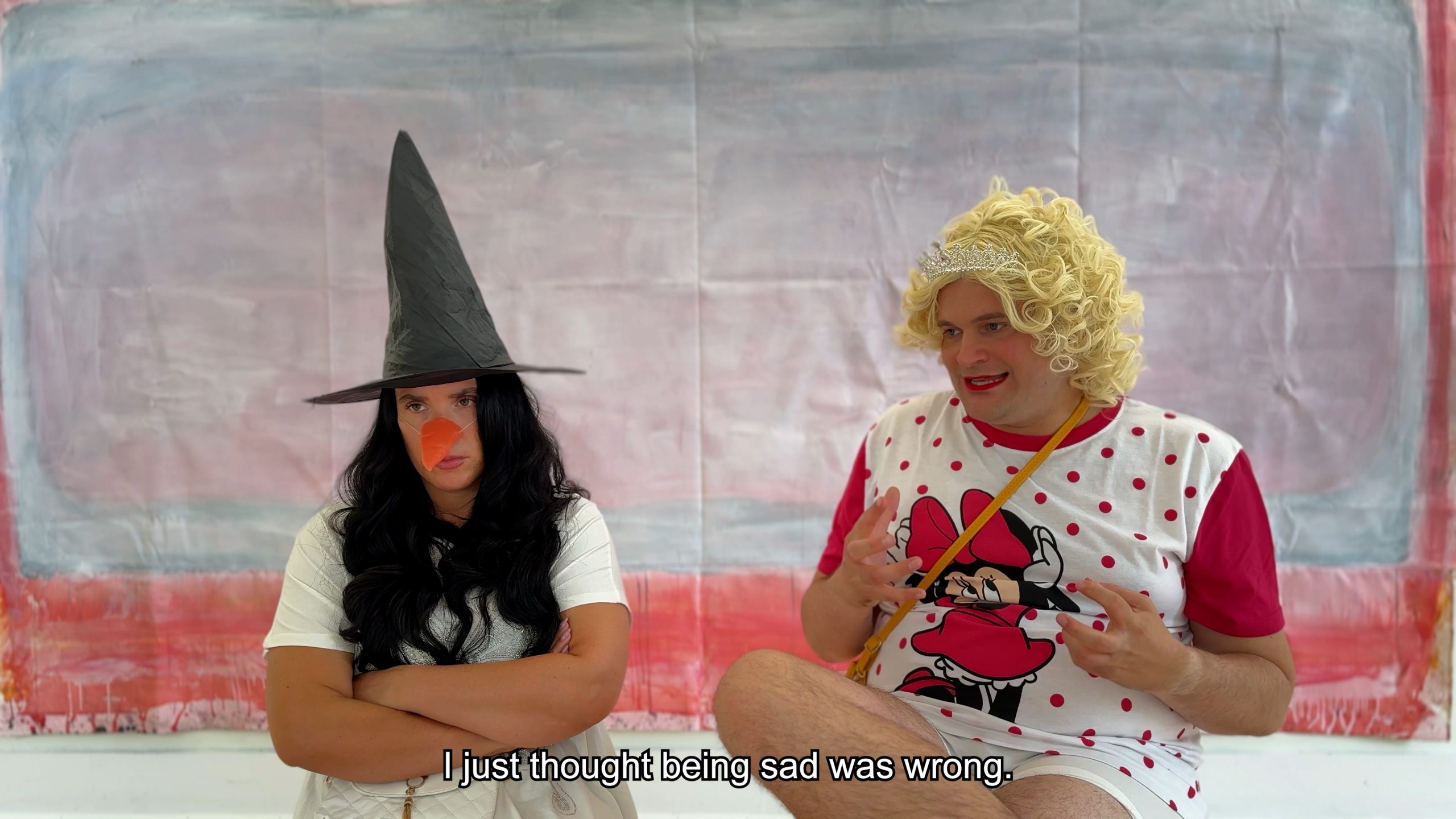 Two people sit in front of a painted backdrop. One is wearing a fancy dress witches hat and carrot nose, they have their arms folded and look fed up. The other is wearing a Minnie Mouse t-shirt, blonde wig, and tiara. They wear red lipstick and are gesturing with their hands. The subtitled caption reads 'I just thought being sad was wrong.'