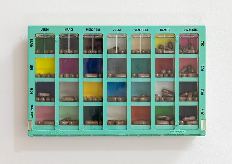 A pastel green pillbox hangs on a white gallery wall. The days of the week are written in French. In each compartment sits brown coloured pills, set against multicoloured backgrounds.