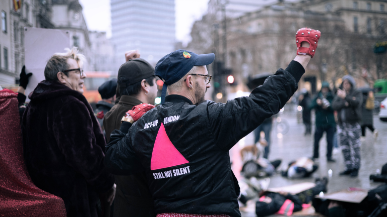 A man in a black jacket raises a fist in an urban space. His wears pink gloves, and on the back of his jacket is a pink triangle with the words ACT-UP London, Still Not Silent embroidered across it. His other hand is on the shoulder of someone with their back to the camera, who also stands alongside an older man.