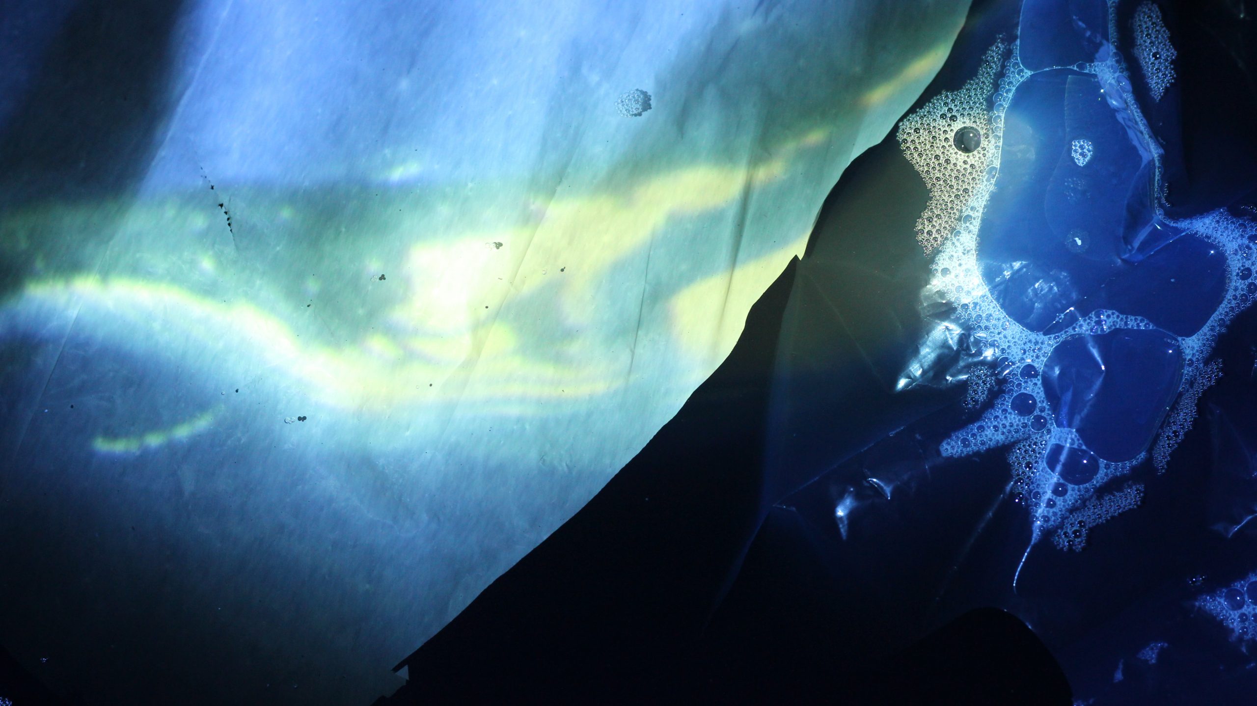 An abstract image shows blues and greens of underwater. A plastic sheet is to the right of the image, something bubbles up against it. A white sheet of plastic covers the left side of the image. Light plays across it.
