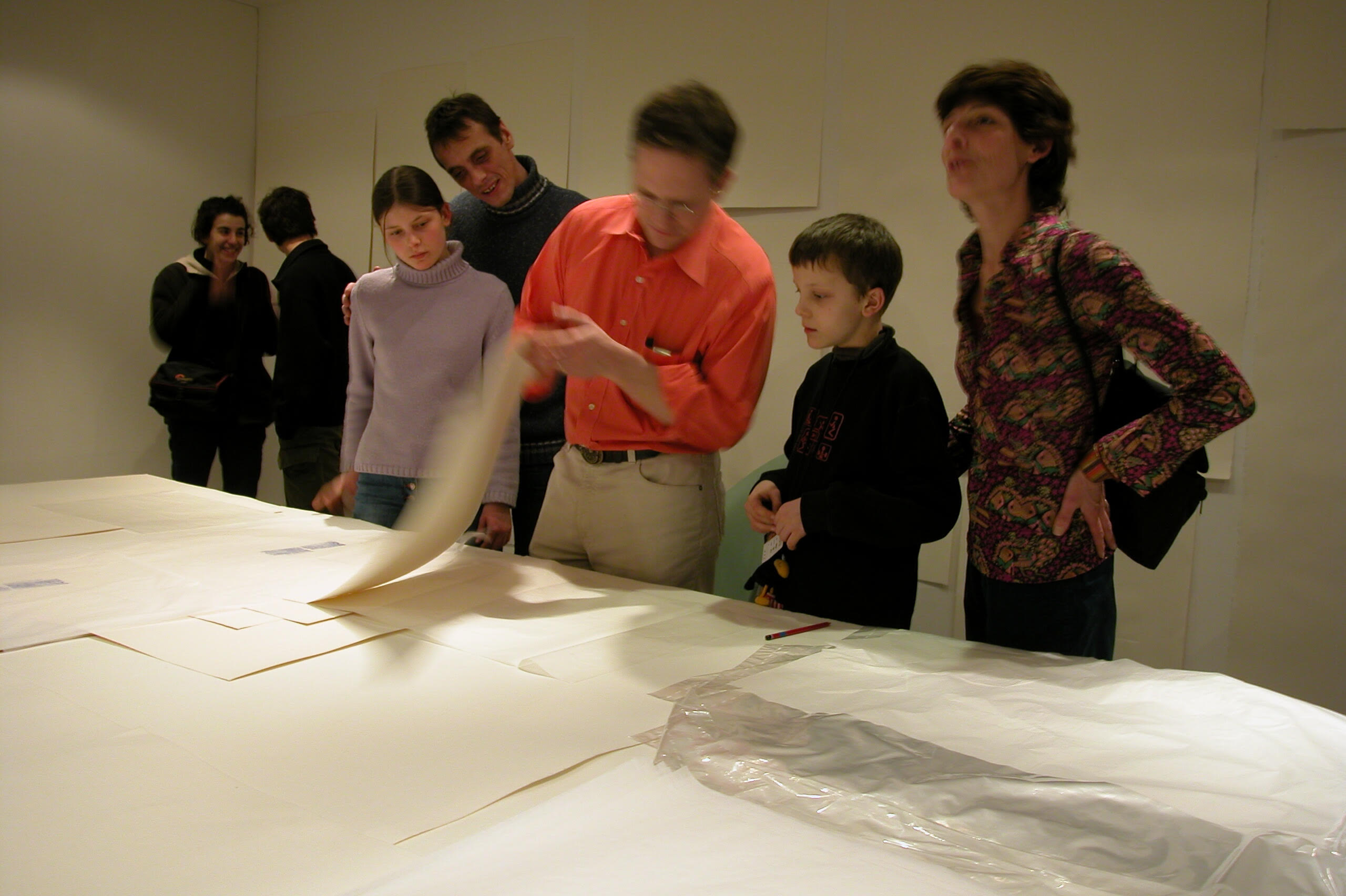 A group of people stands around a large table examining sheets of paper in a well-lit gallery space.