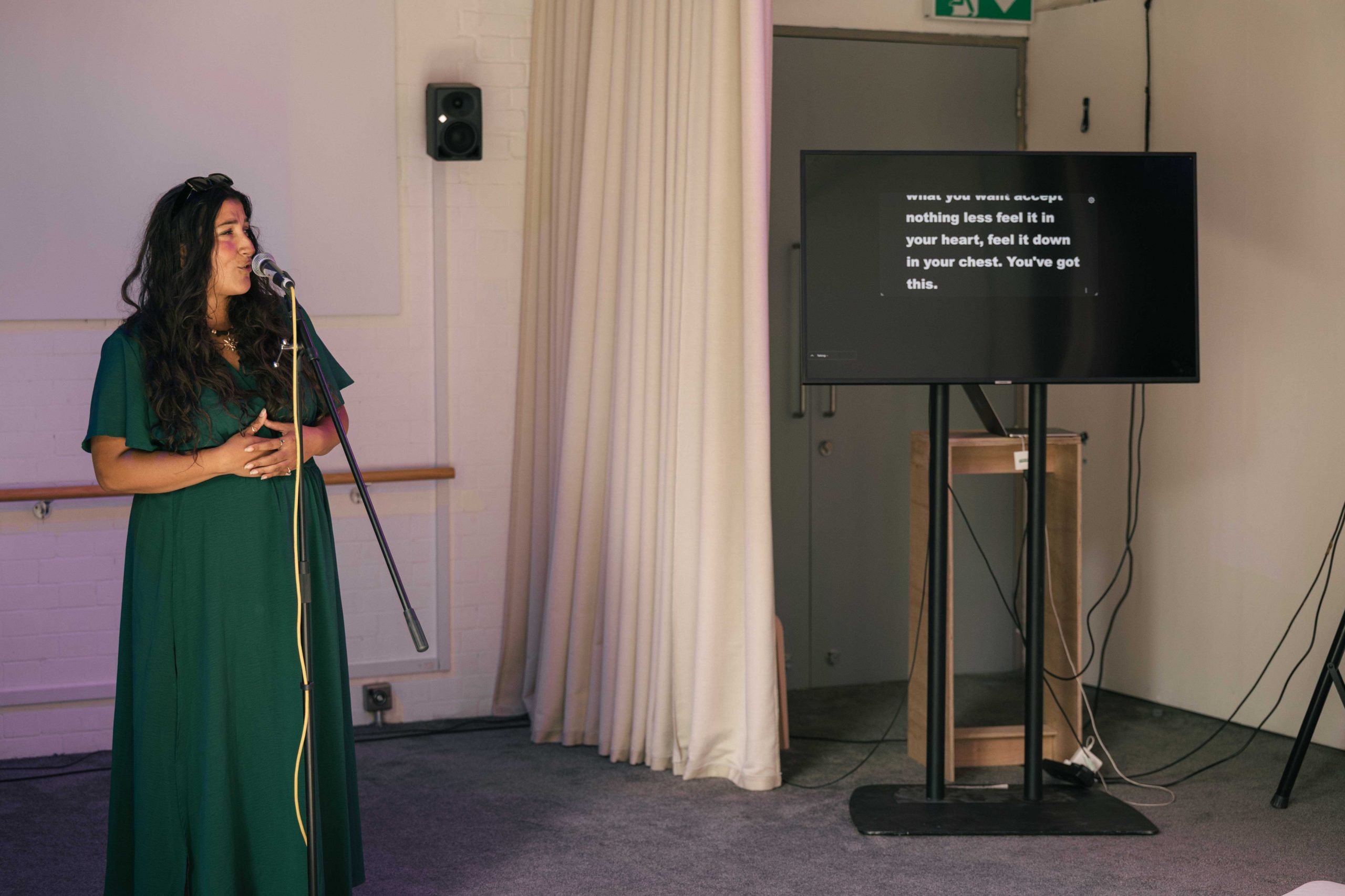 a woman standing behind a microphone. in the background to left is a television screen on a high metal stand displaying what she is saying.