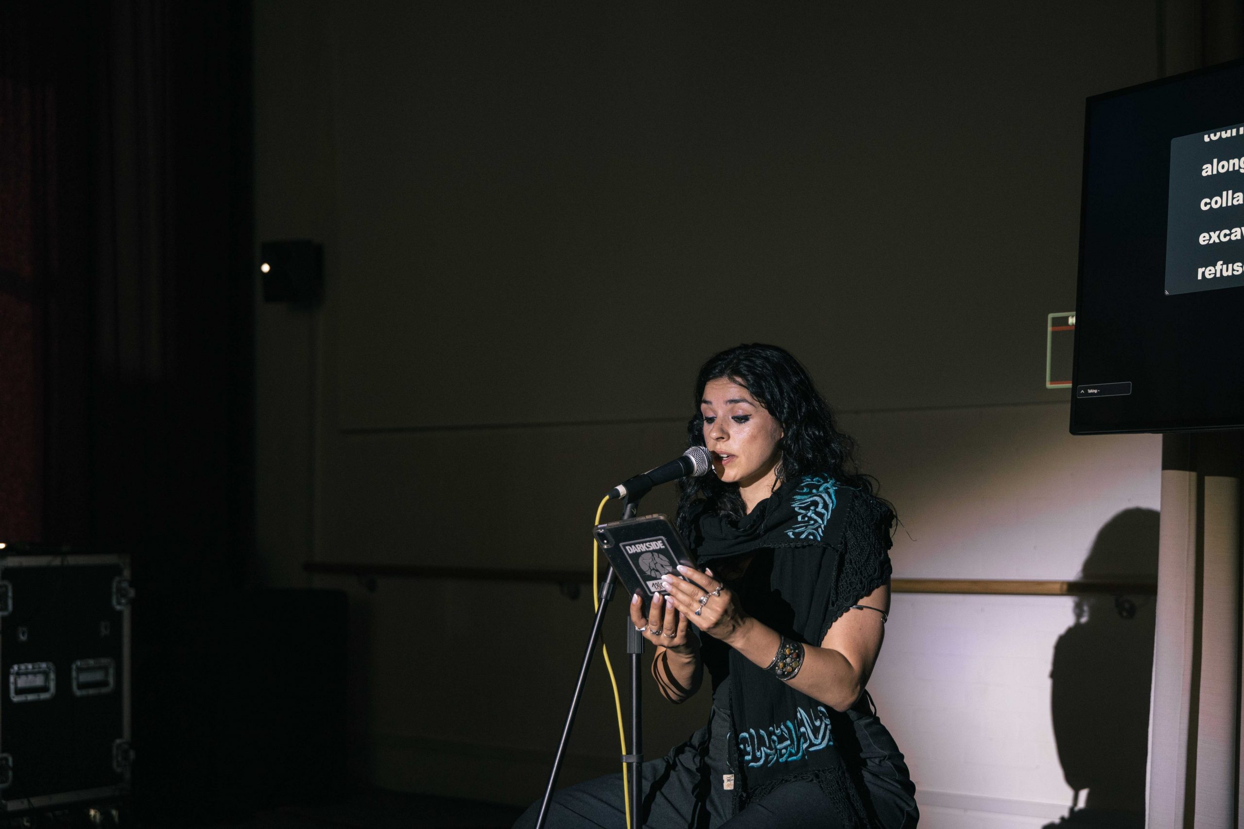 A woman with black hair sits reading into a microphone under a spotlight.