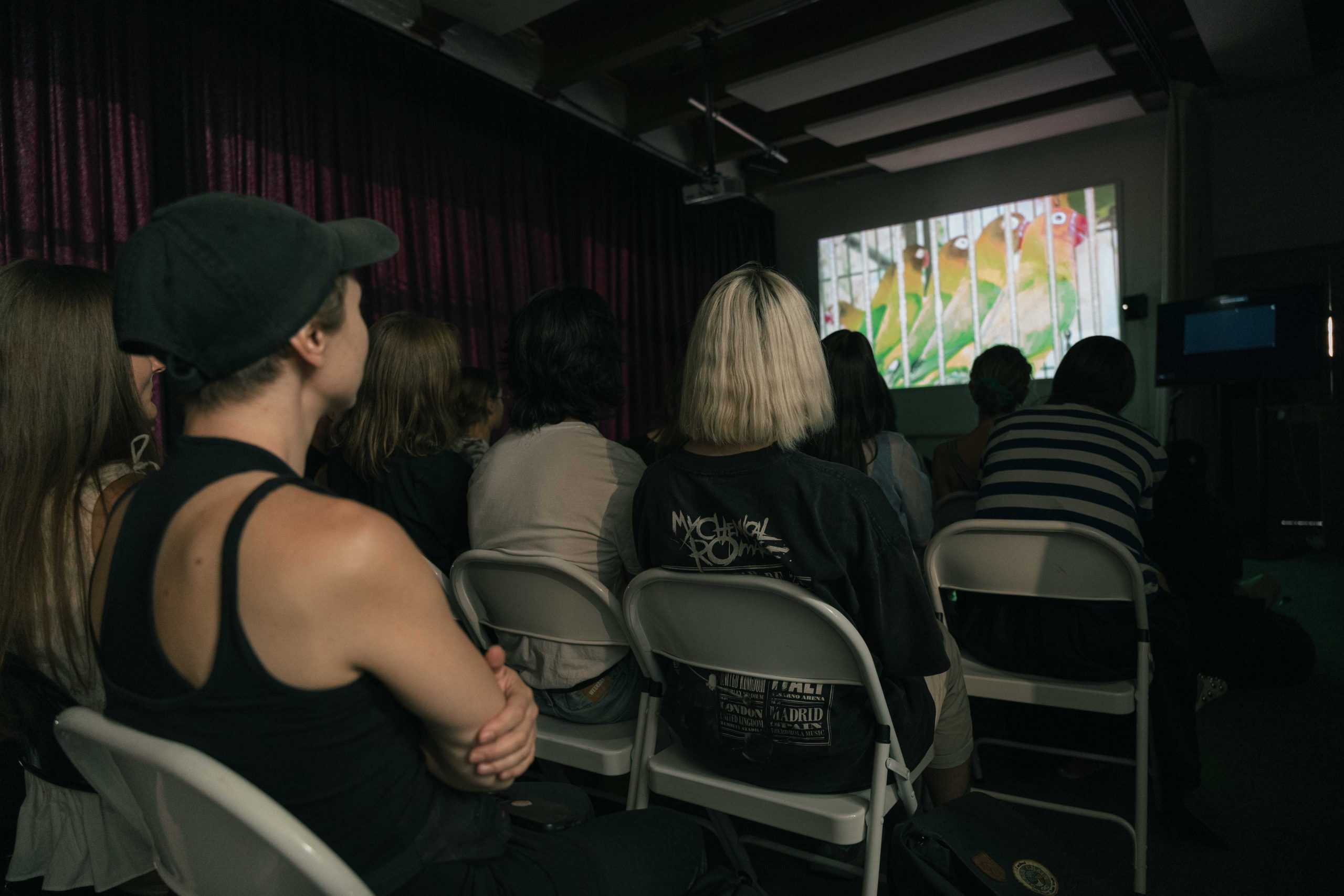A group of people sitting on white metal chairs look at a screen in front.