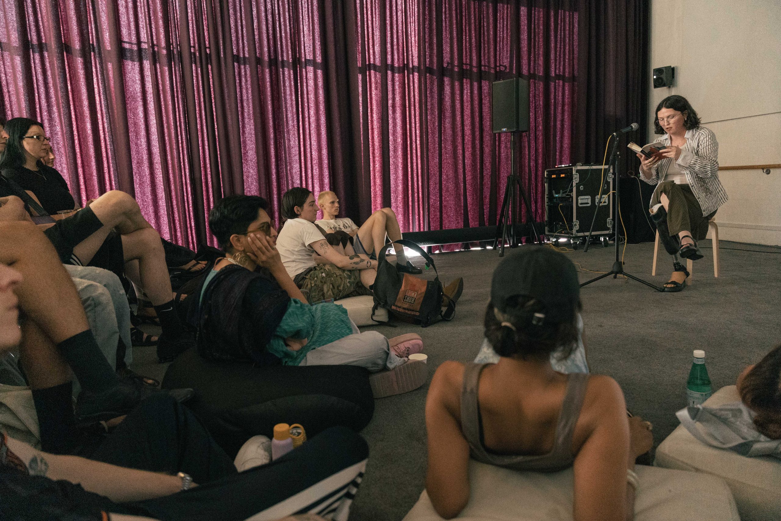 A woman sits reading into a microphone in front of a group of people lying on beanbags and sitting on chairs. A purple curtain hangs across the background.