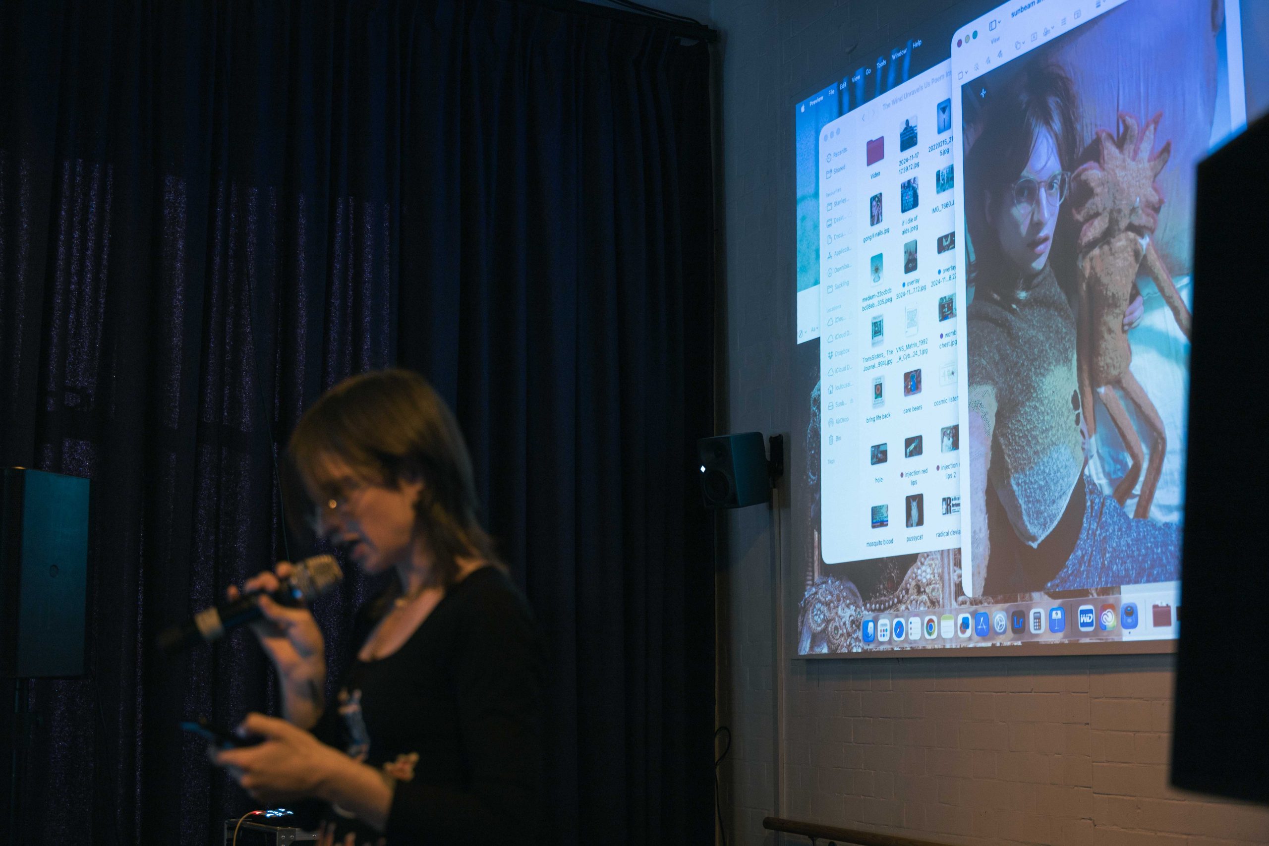 A woman reads into a microphone, a computer screen is projected behind her.